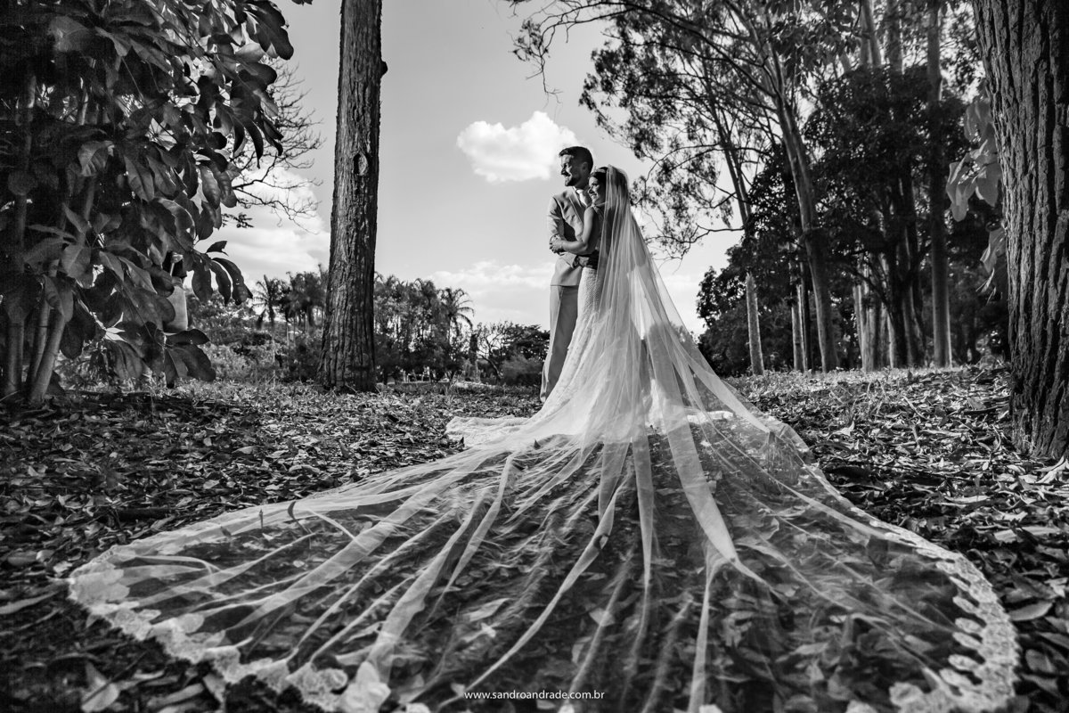 Uma linda fotografia da noiva de costas pegando todo o vestido da la fiancée e um lindo véu longo, Michel e Luiza estão abraçados, olhando lateral, mais uma belíssima criação de Sandro Andrade fotógrafo de Brasilia.