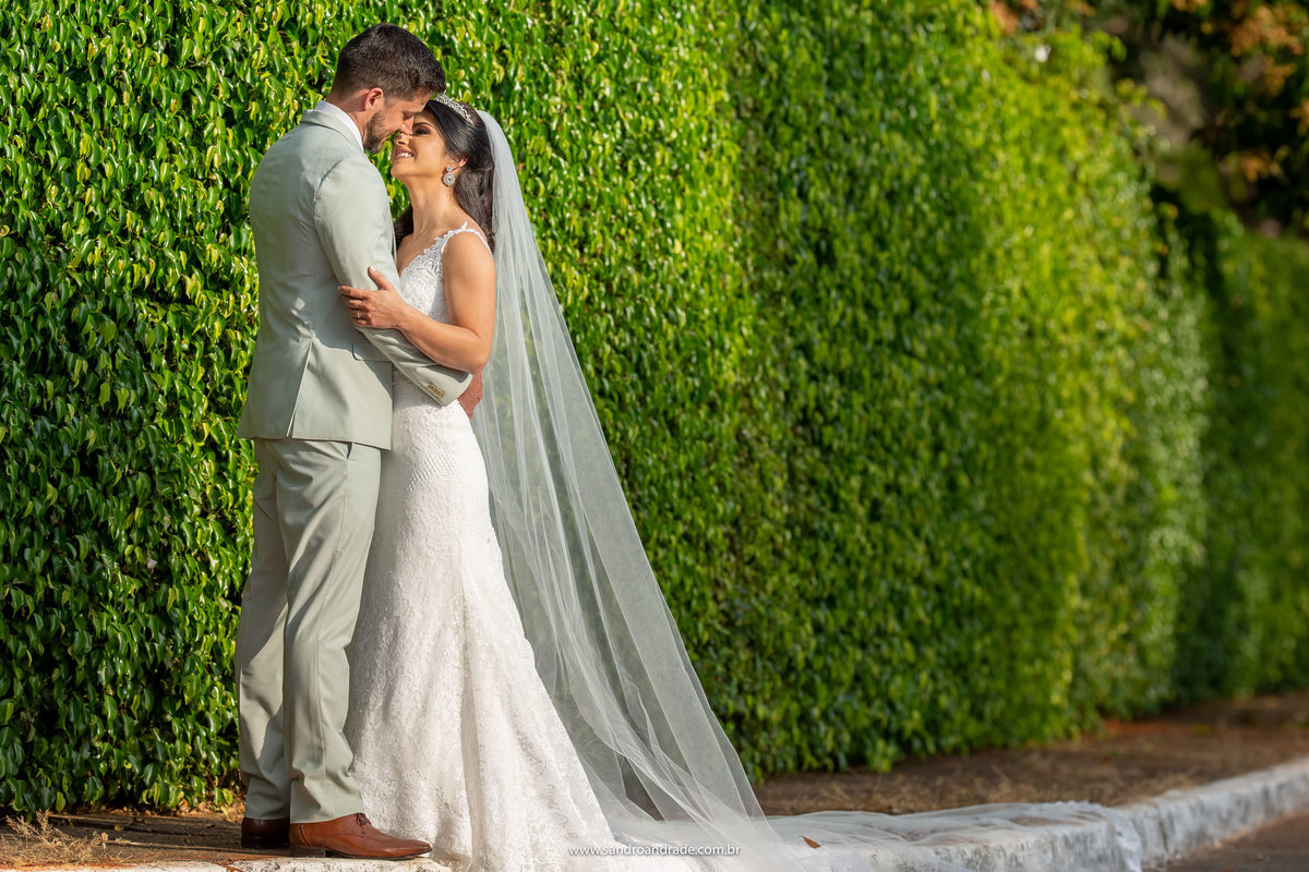 O casal está de frente um para outro nesta fotografia colorida, ainda na Península, sorrindo e apaixonados eles se olham, muito verde de um muro com folhas ao fundo desta bela imagem.