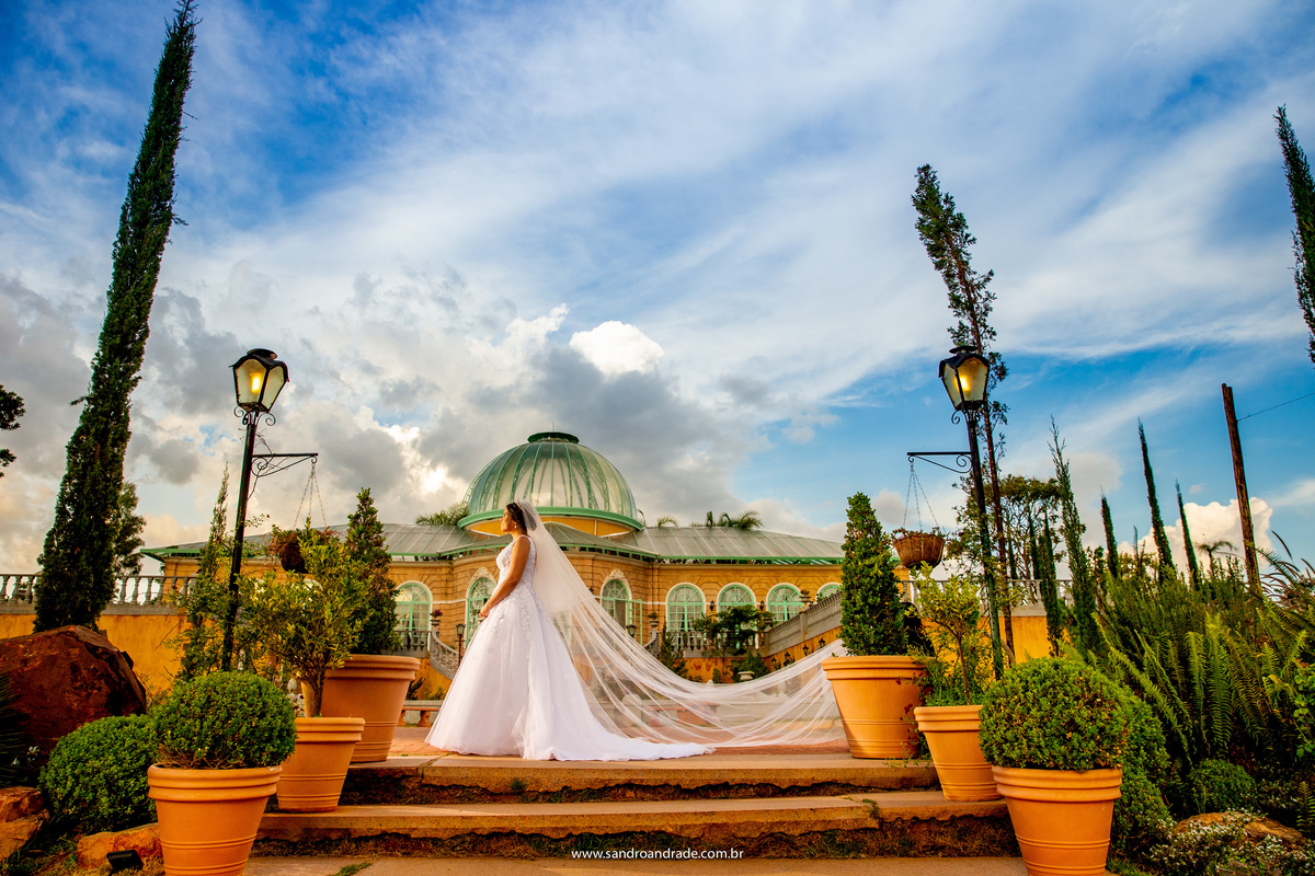 No giardino da Villa, pegando todo o palazzo no fundo em tons alaranjados e o contraste do verde do jardim e o céu azul com muitas nuvens, Julia está linda com seu véu voando e uma luz linda no rosto.