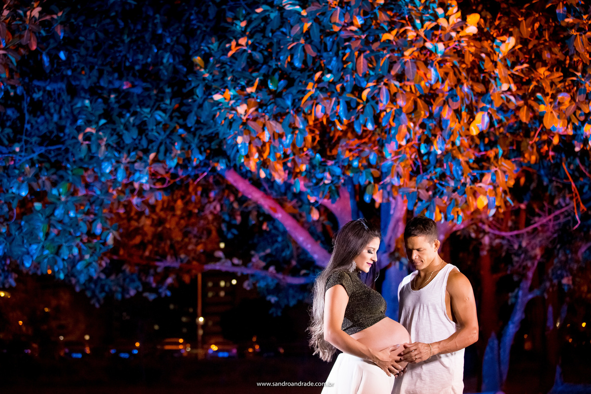 Uma linda fotografia com muitas cores, de frente um para o outro e olhando para a barriga, Sandro brinca com cores nesta imagem, em tons de azul, roxo e laranja ele ilumina a arvore que está atras do casal.