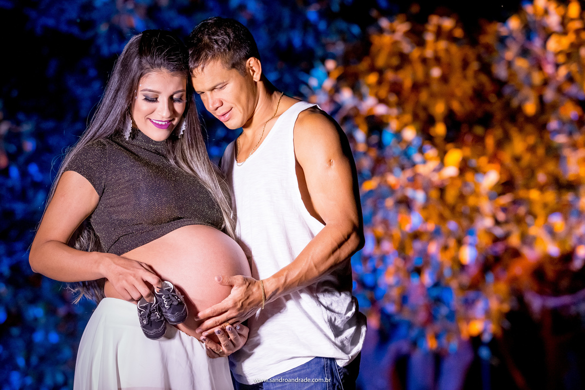 Retrato do casal, abraçados, ele segura na barriga e sua esposa está com o sapatinho do bebê, um menino, os dois estão olhando para o sapatinho, cores e seus amores, linda e perfeita combinação.