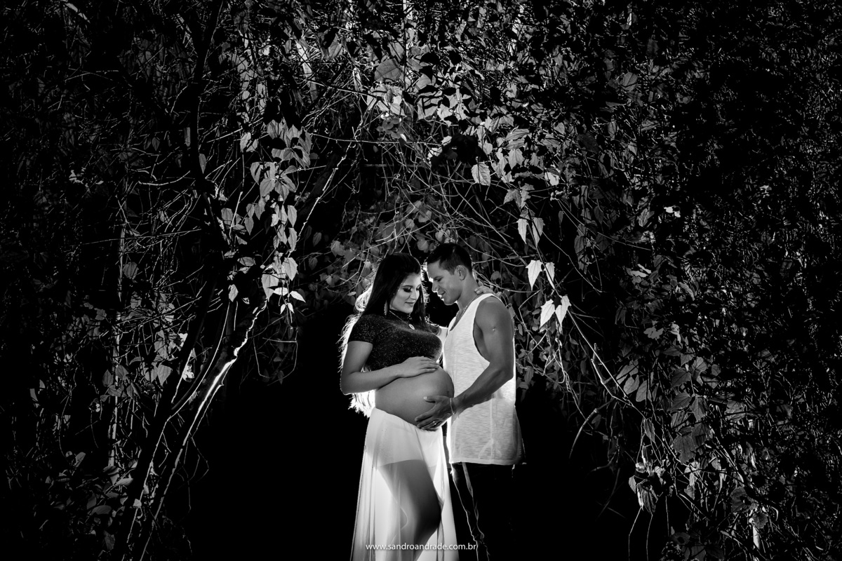 Preto e branco é sempre um arraso, o casal está bem juntinhos no centro da fotografia em baixo de uma arvore formando o arco, abraçados e olhando para a barriga eles estão felizes pela chegada do fruto deste amor.