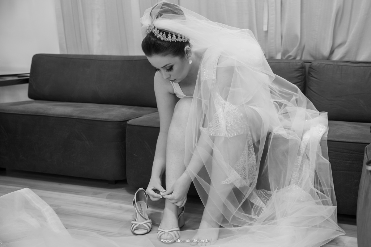 Ela já está se arrumando, já retirou seu roupão e agora está colocando o sandália que irá usar com seu lindo vestido. Uma fotografia clássica da noiva em preto e branco.