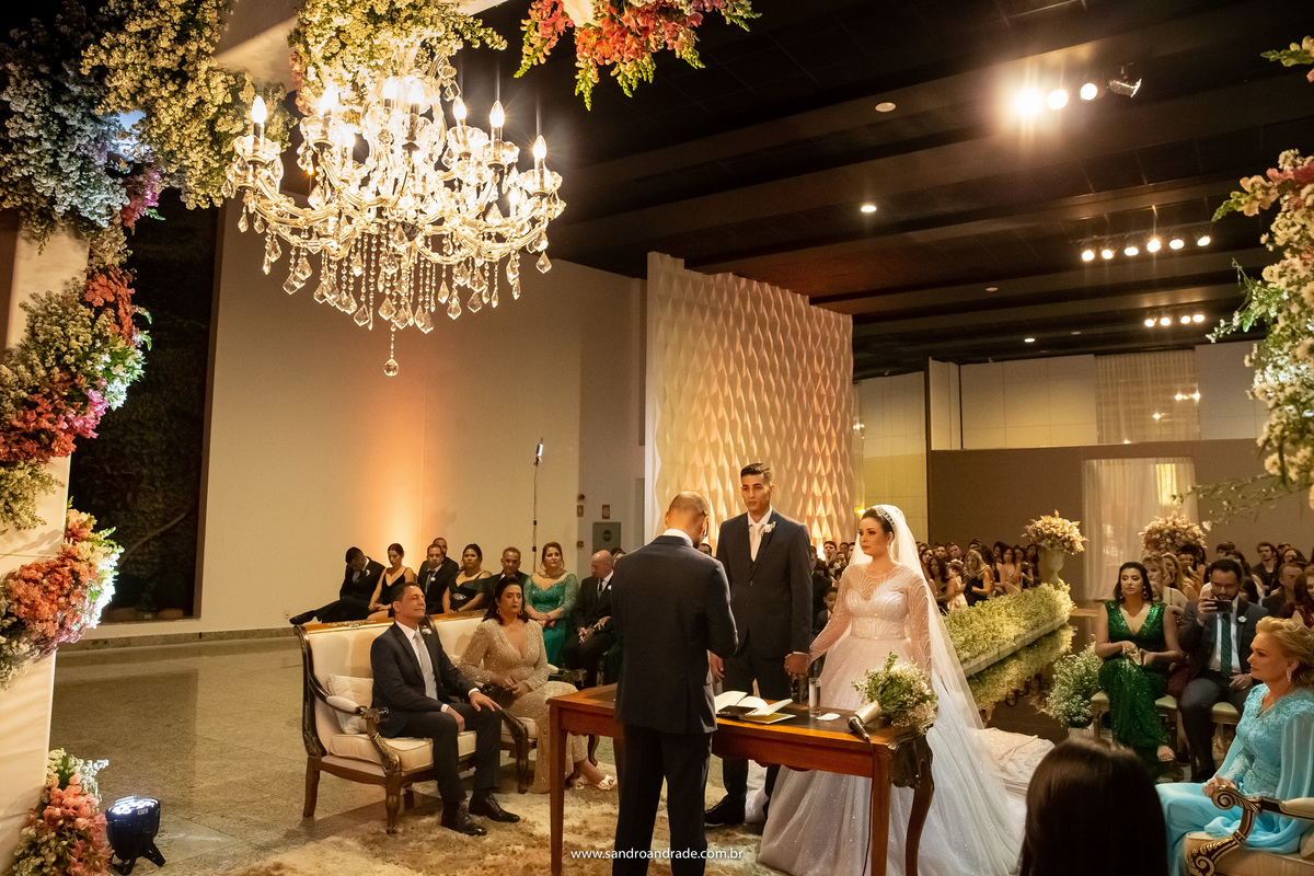 Uma imagem mostrando a linda decoração com um lustre maravilhoso, o casal a frente do pastor que realizou esta linda cerimonia e os pais nas laterais.