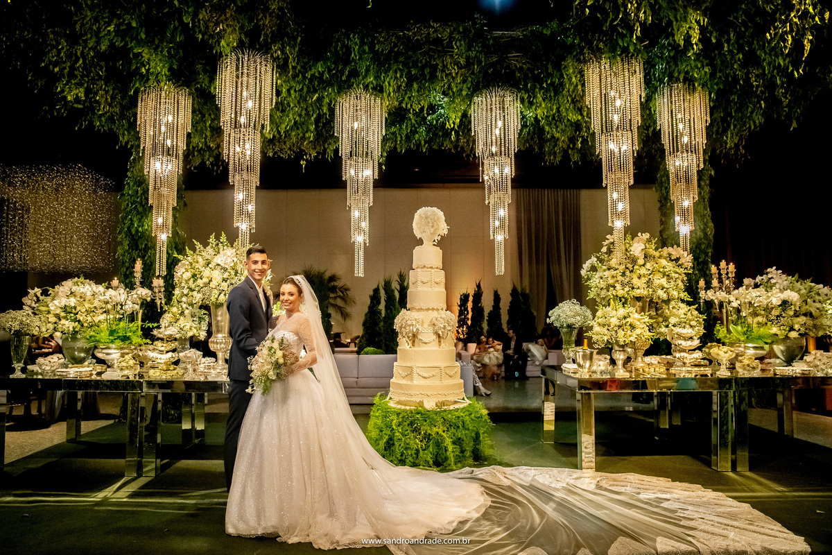 O casal está posando para a fotografia na frente da mesa do bolo, ela com seu belíssimo vestido de noiva do ateliêr Fernando Peixoto e ele com seu terno da máxime noivos e black tie.