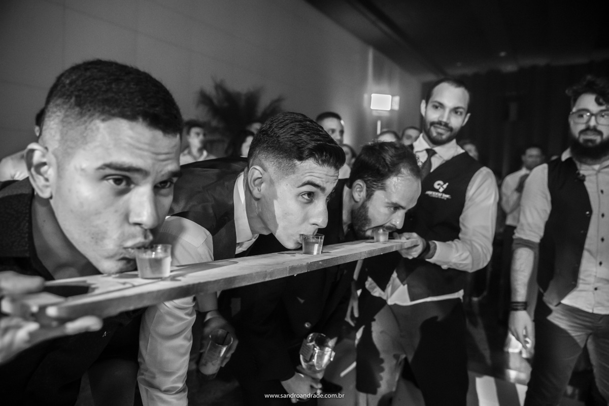 O noivo com mais dois amigos tomando tequila em uma bandeja suspensa, onde os três tomam a bebida de uma vez e em uma só golada.
