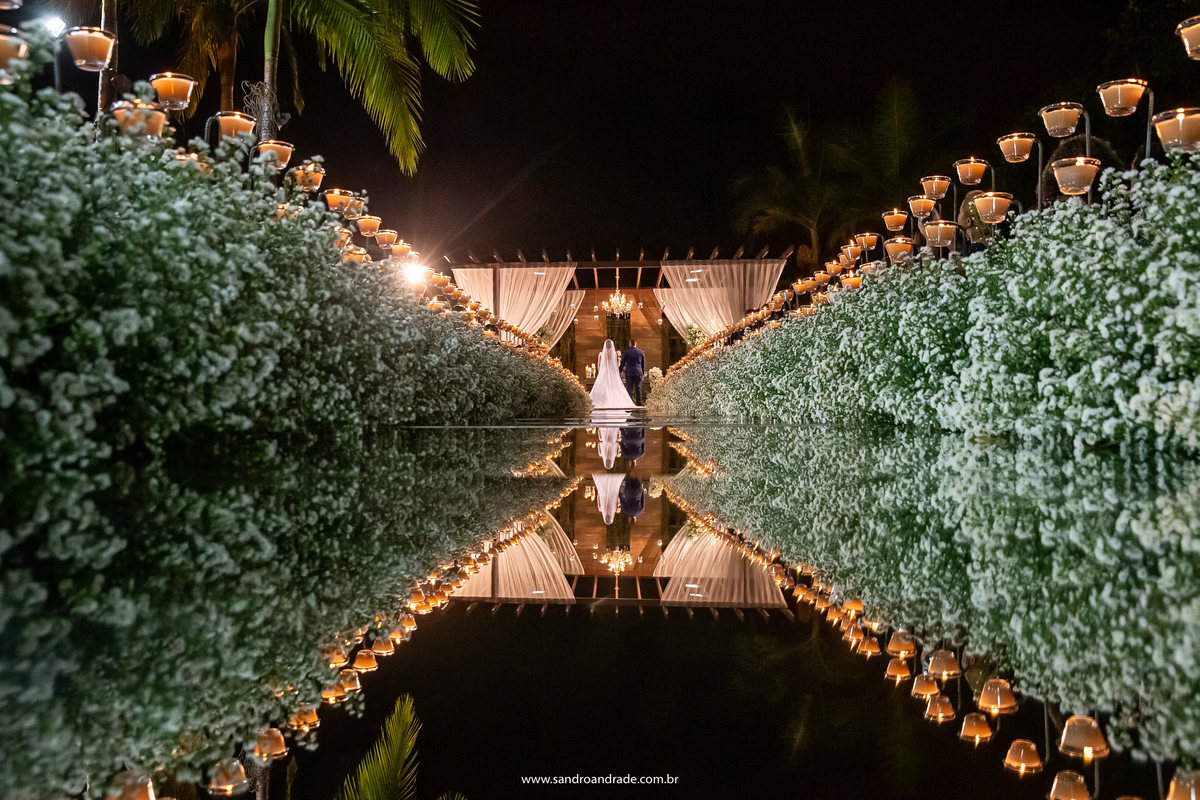 Uma linda fotografia do nosso artista Sandro Andrade durante o cerimonia, o casal esta de costas no altar e ele pega todo o caminho com as velas, as flores e o tapete espelhado, Sandro é um dos melhores fotógrafos de casamento de BSB.