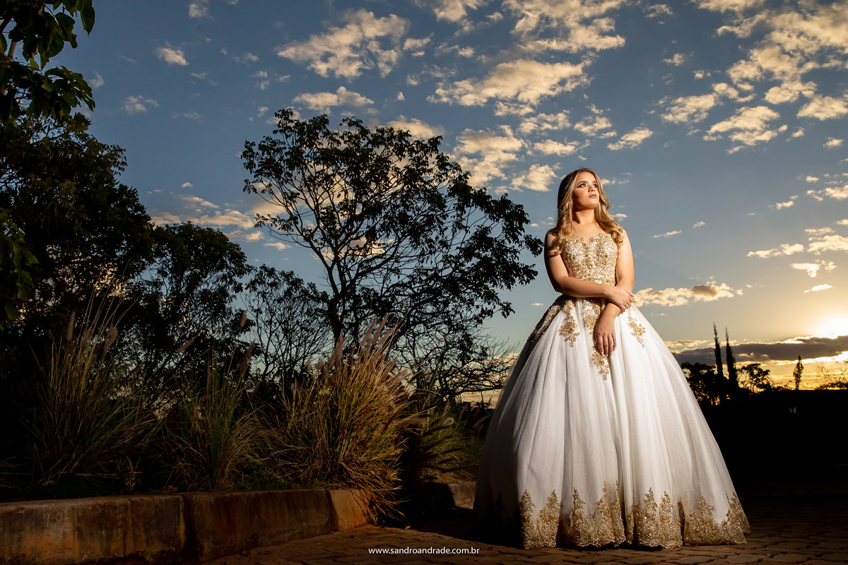 Uma fotografia de baixo, com Amanda de corpo inteiro e um lindo céu azul e do lado esquerdo as árvores da mata.