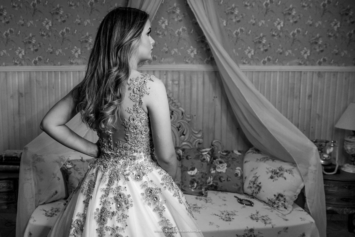 Em pé e de costas, dentro do quarto com seus lindos cabelos soltos e a mão na cintura.