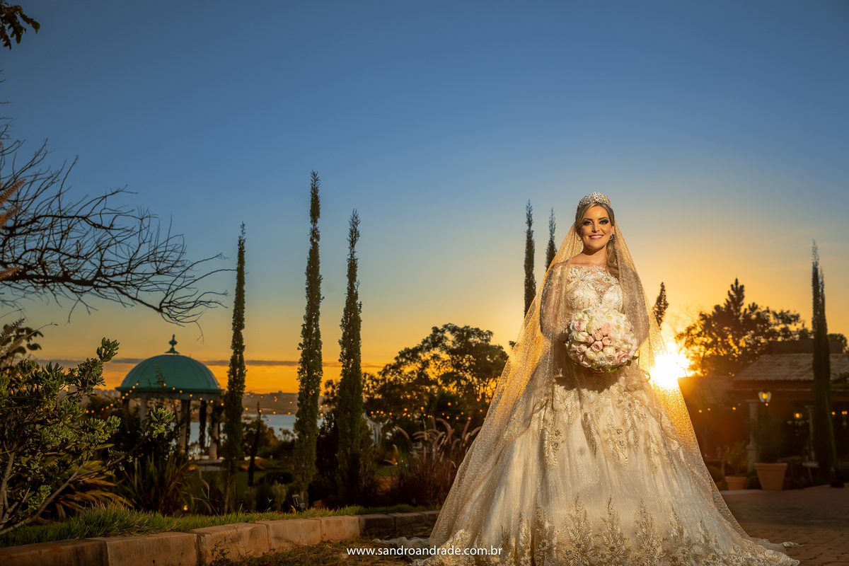 Cassia está com seu lindo buque, no canto direito da fotografia, o sol se pondo atras dela e todo o jardim da área do gazebo ao fundo.