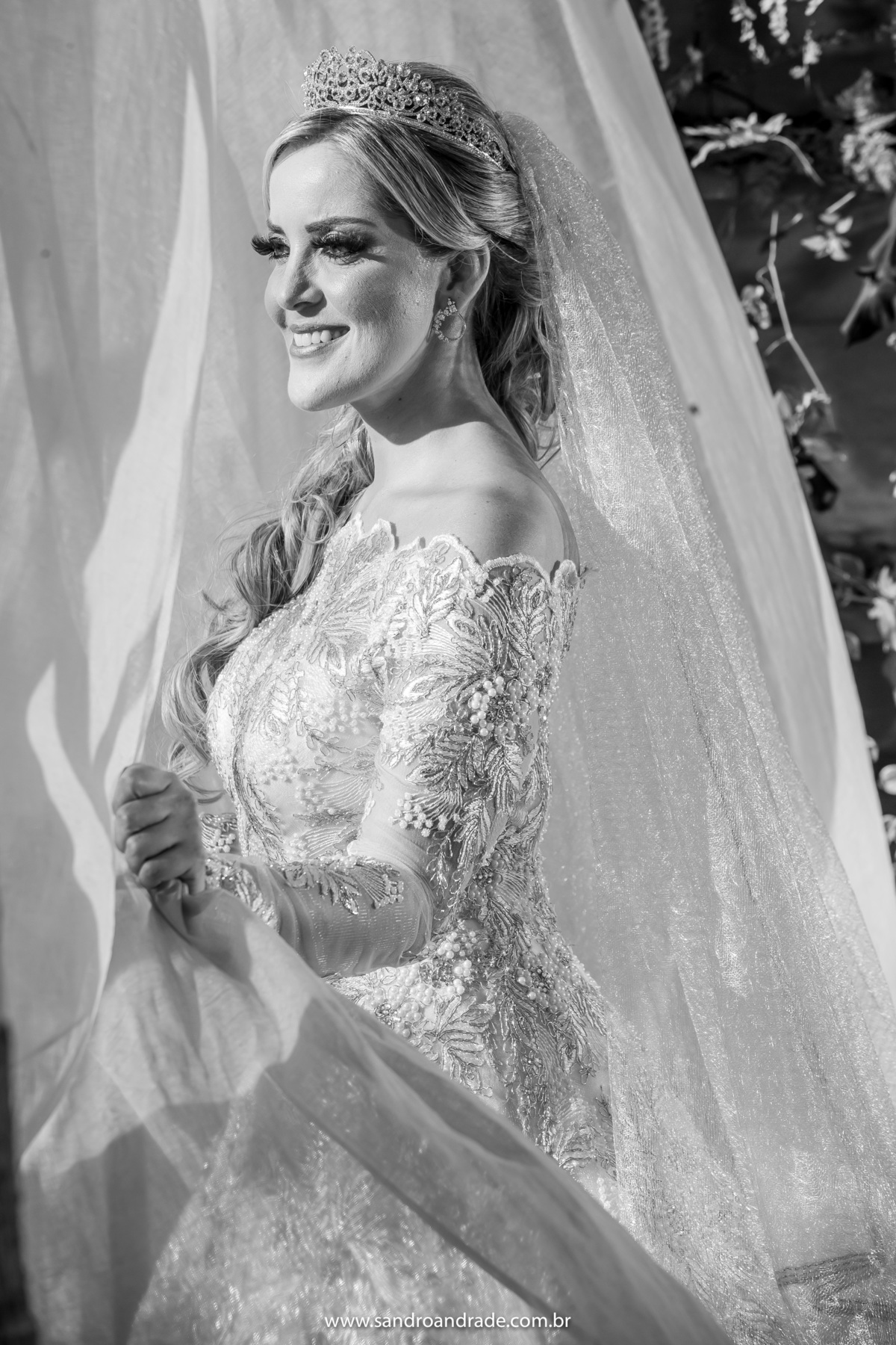 A noiva em preto e branco, segurando a cortina e deixando a luz do sol iluminar seu rosto.