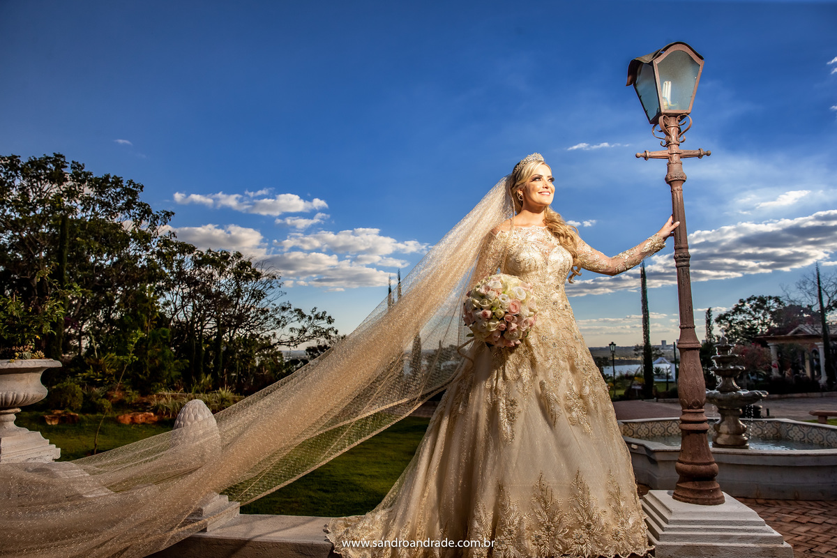 Em cima de um muro e ao lado de um poste de luz, Sandro Andrade usa luz artificial para iluminar a noiva e pegar esse lindo azul do céu, linda foto com o véu voando.