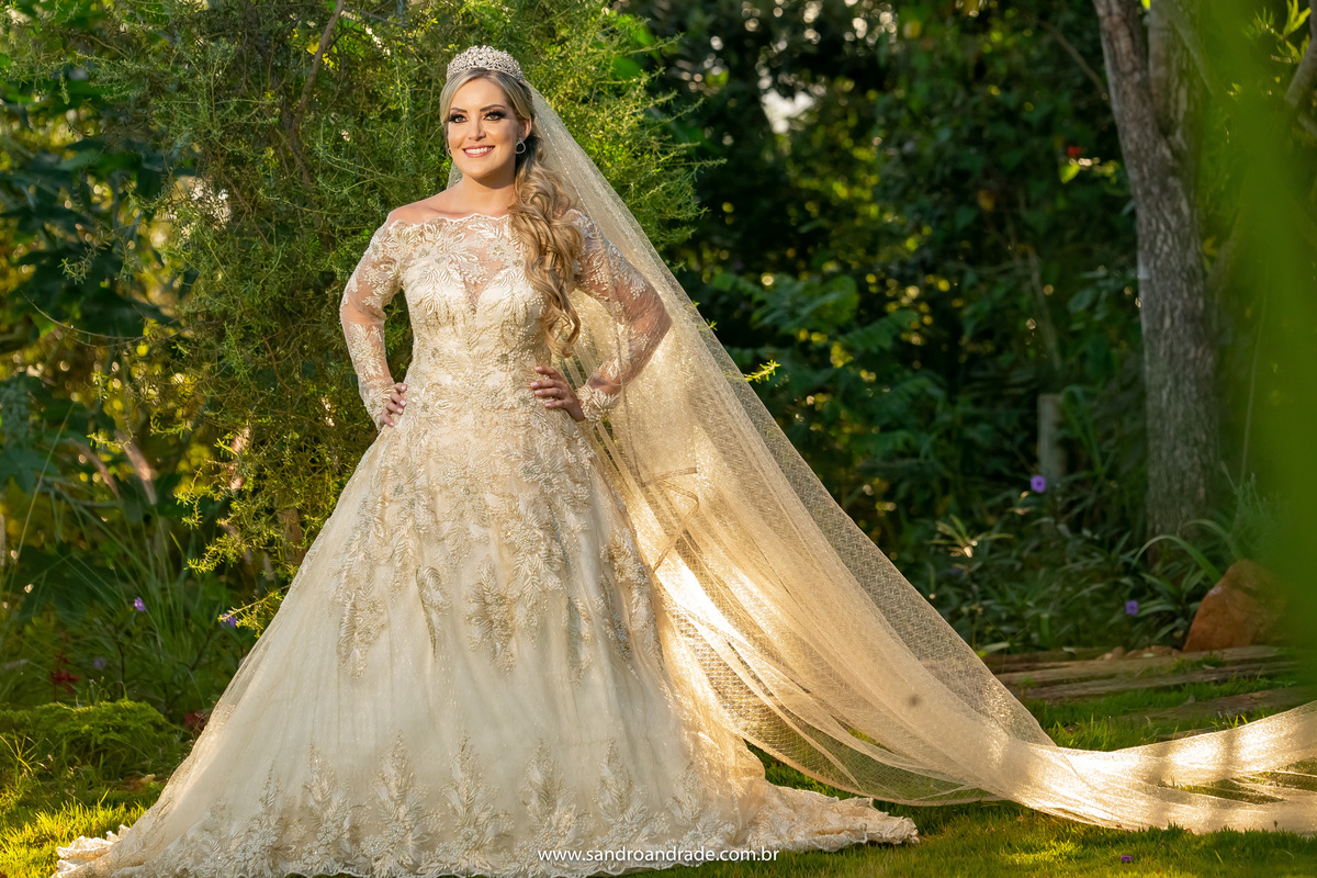 Uma fotografia da noiva de corpo inteiro no jardim da Villa, com as mãos na cintura e véu todo arrumado, ela toda de dourado em contraste com o verde das árvores.