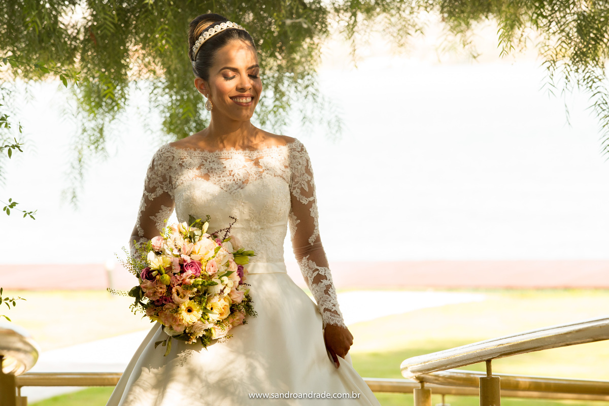 Sorrindo e de olhos fechados, Taísa está com seu lindo buque de noiva na mão, numa ponte que tem no Porto Vitória, Sandro Andrade fotografo de casamentos em Brasília faz esta bela imagem com o lago de fundo.