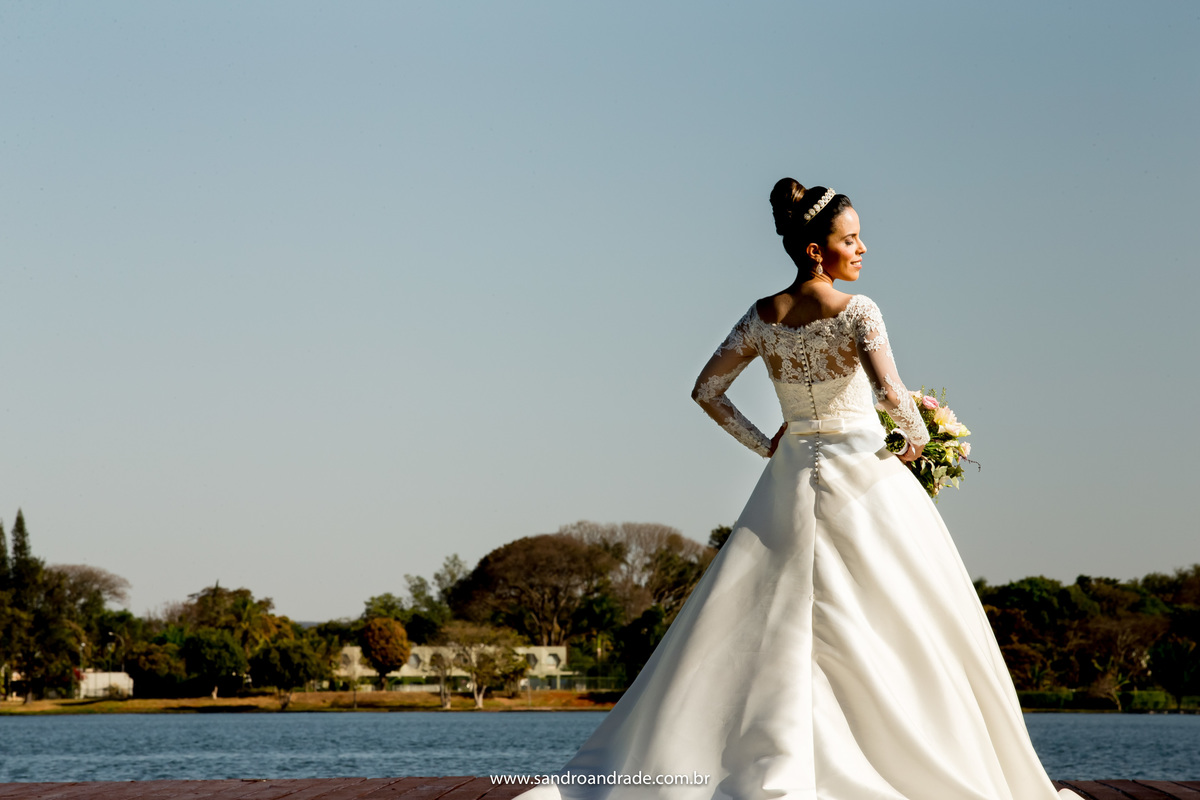 Na beira do lago, de costas, segurando seu buque, sem véu, uma linda imagem colorida.