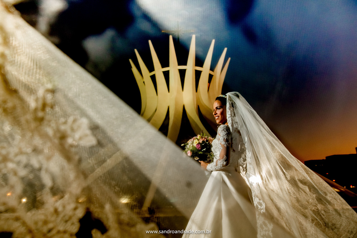 Taísa olha de perfil, enquanto Sandro Andrade faz um lindo retrato dela com o véu voando na frente da lente.