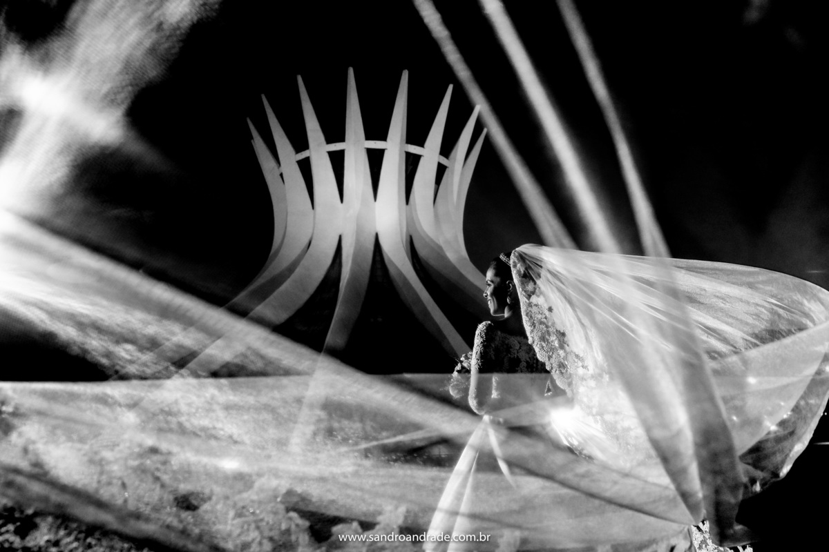 Uma imagem PB, da noiva de costas, sorrindo e o véu voando na frente das lentes de Sandro Andrade.