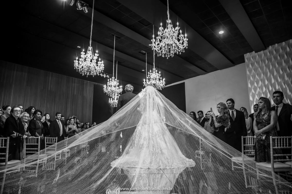 Fotografia preto e branco da noiva entrando de costas, mostrando os convidados a admirando e to seu lindo véu aberto, fotografia artística por Sandro Andrade o mago da luz.