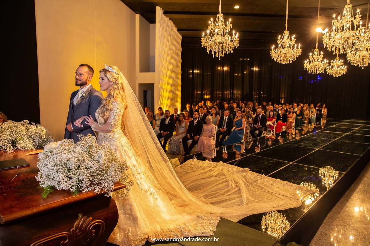 Os noivos de corpo inteiro no altar e todos os lustres e convidados ao fundo.