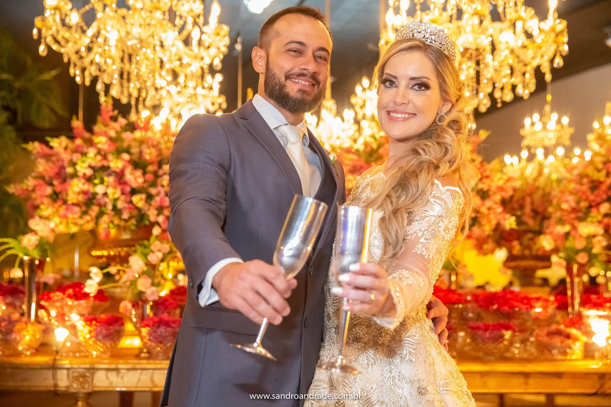 Os noivos estão na mesa do bolo, fazendo o brinde, este lindo retrato dos noivos é colorido, cheio de vida e alegria.