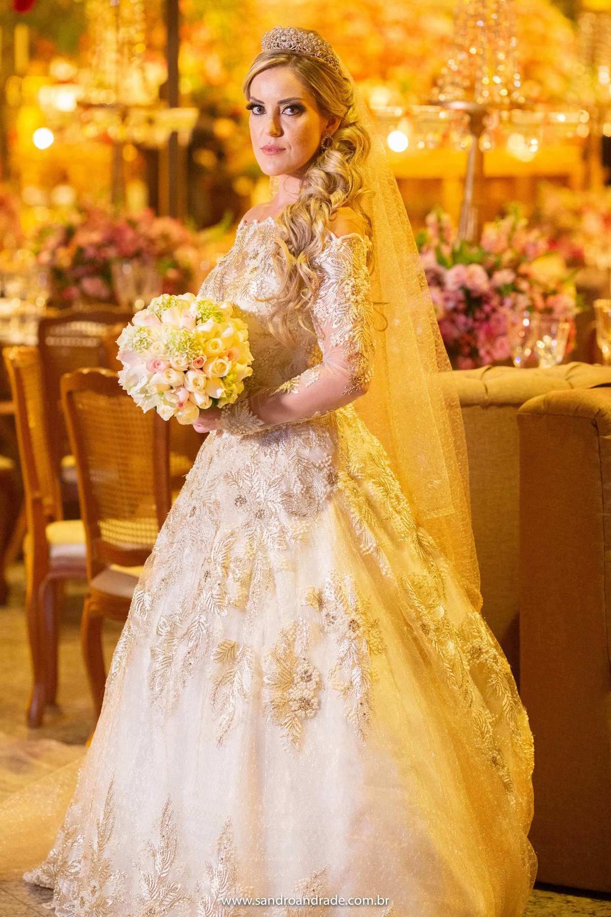 Uma fotografia da noiva de corpo inteiro em meio a decoração da festa com seu lindo buque na mão, tudo em tons de amarelo e dourado assim como o vestido da noiva.