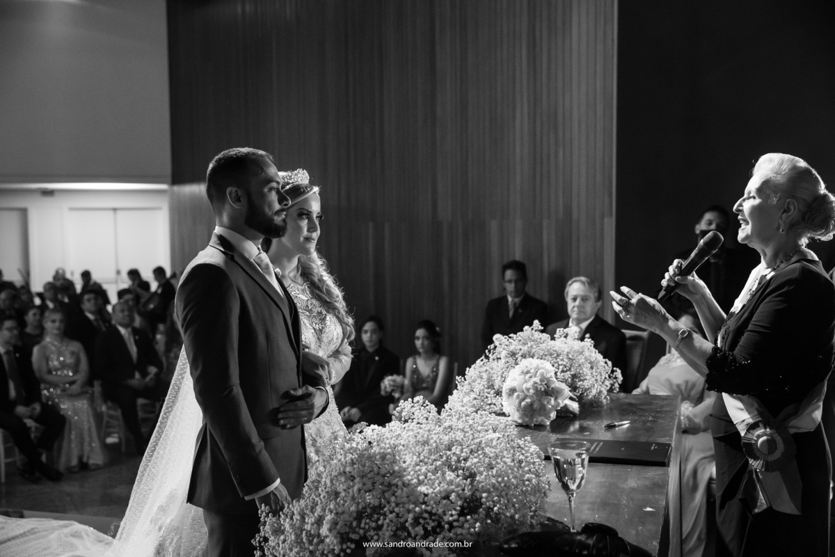 Uma fotografia preto e branco, lateral do altar, Cassia e Rafael um ao lado do outro e a dra Eunice celebrando o casamento.