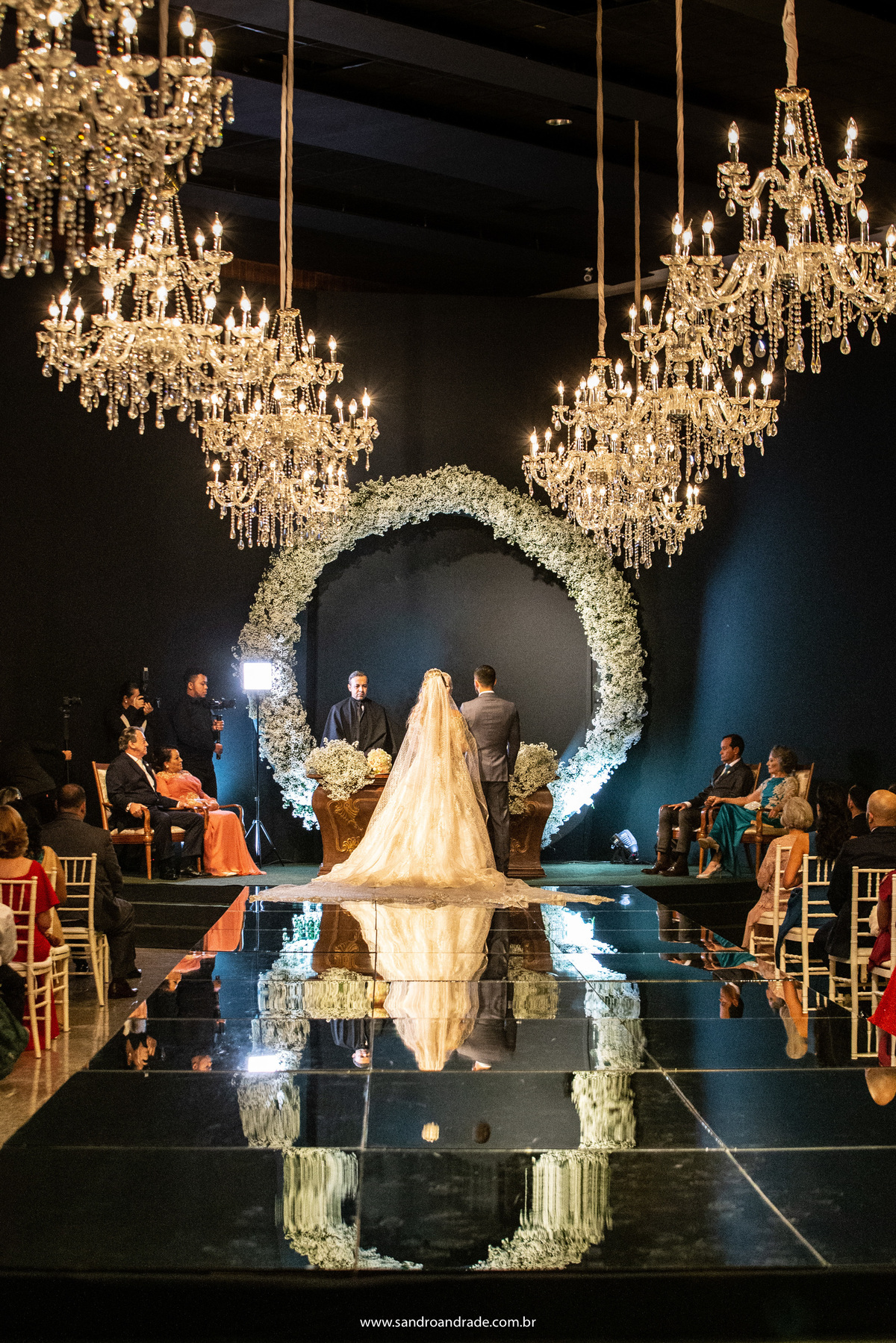 O casal de costas no altar, mostrando os lustres, o arco de flores sorriso de Maria e o reflexo no tapete espelhado.