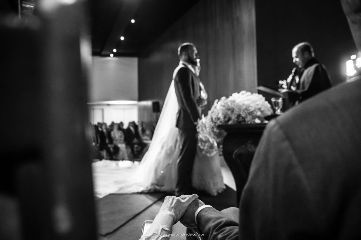 Enquanto a cerimonia é realizada, Cassia e Rafael estão ao fundo e os pais estão servindo de moldura nesta fotografia preto e branco, os pais de mãos dada e em destaque a aliança da mãe do noivo.