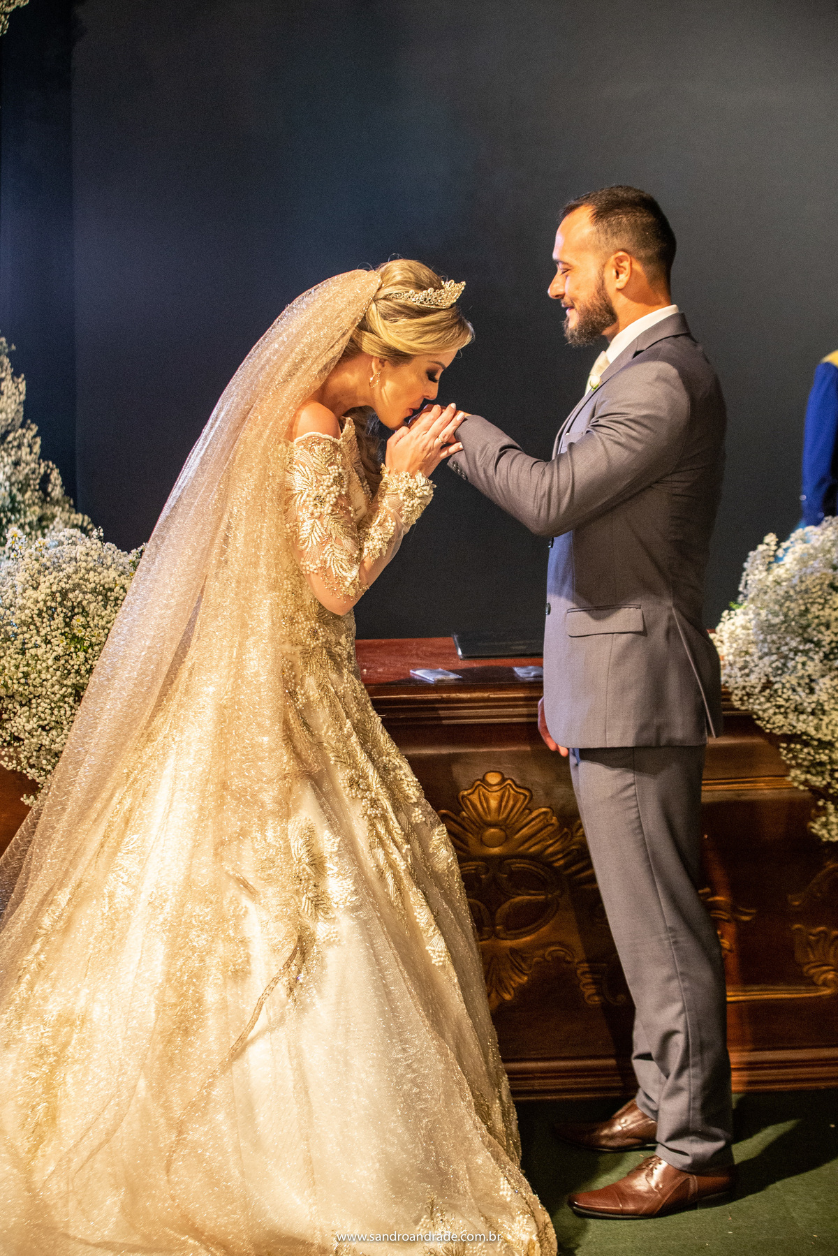 Uma fotografia vertical, colorida, da noiva beijando a mão de seu noivo.