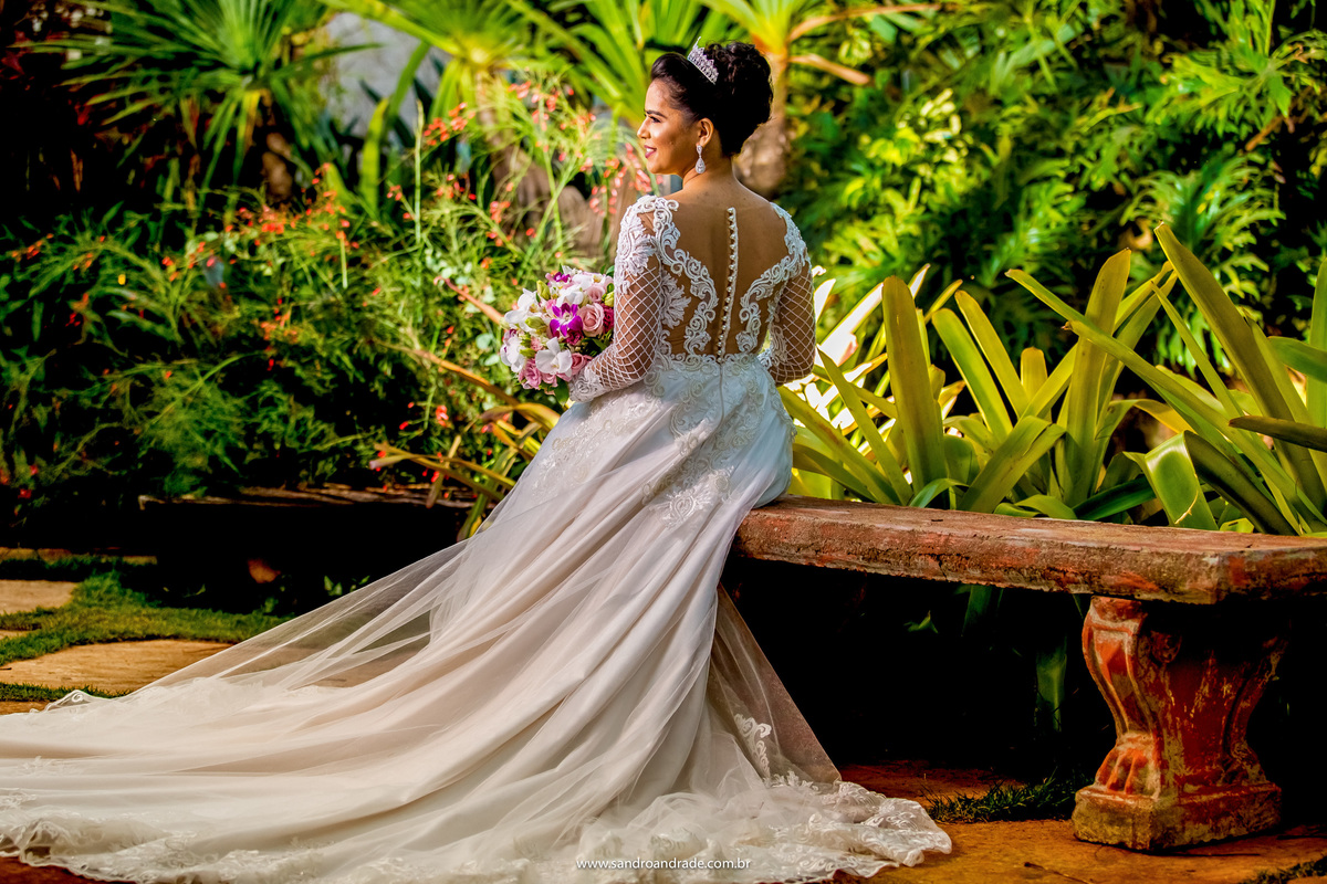 A noiva está de costas, sentada, olhando lateralmente e com o buque na mão, sorri com uma luz linda no rosto e como luz de fundo o sol.