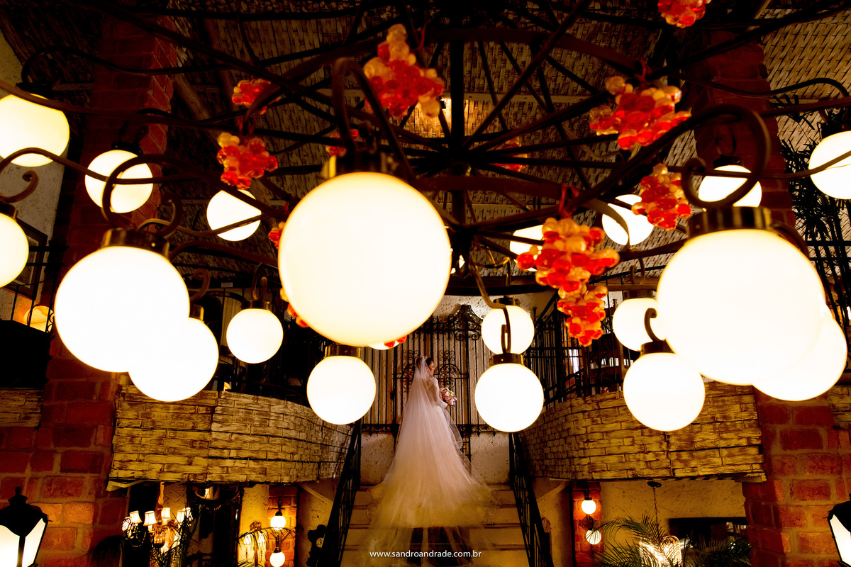 Flavia está no topo da escadaria do Spazio Villa Regia e o lustre esta na frente dela fazendo uma fotografia emoldurada, emoldurando a noiva que está linda e ansiosa, fotografia de Sandro Andrade fotógrafo de casamentos.