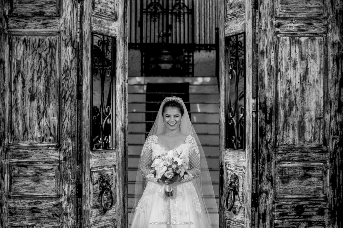 Sorrindo, com seu buque na mão e em preto e branco, uma linda fotografia de Flavia entre as portas lindas de madeira na entrada principal do salão.