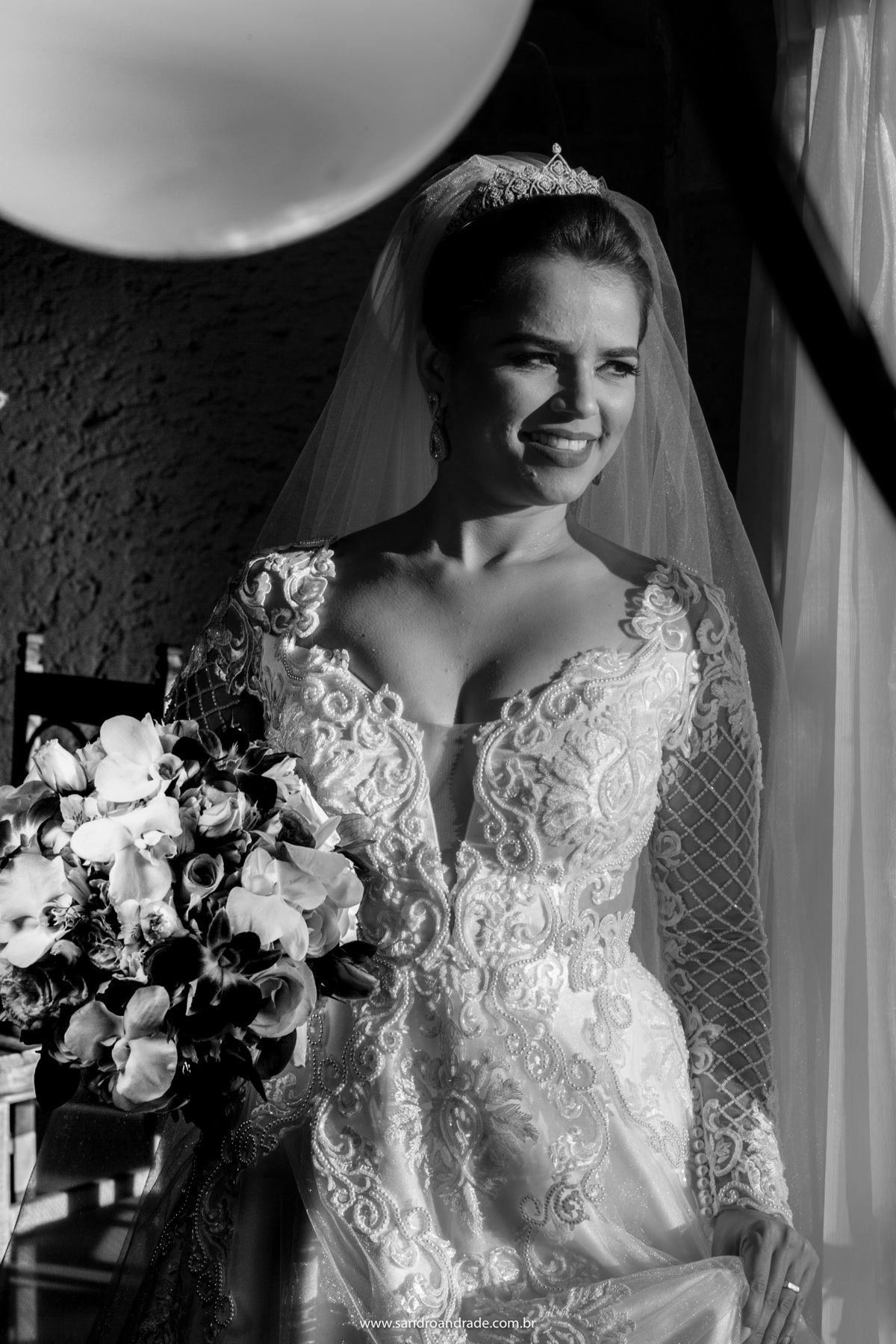 Retrato da noiva, em pé, preto r branco, sorrindo, com o buque em uma das mãos, enquanto a outra segura a saia do vestido, linda imagem feita pelo fotógrafo de BSB Sandro Andrade.