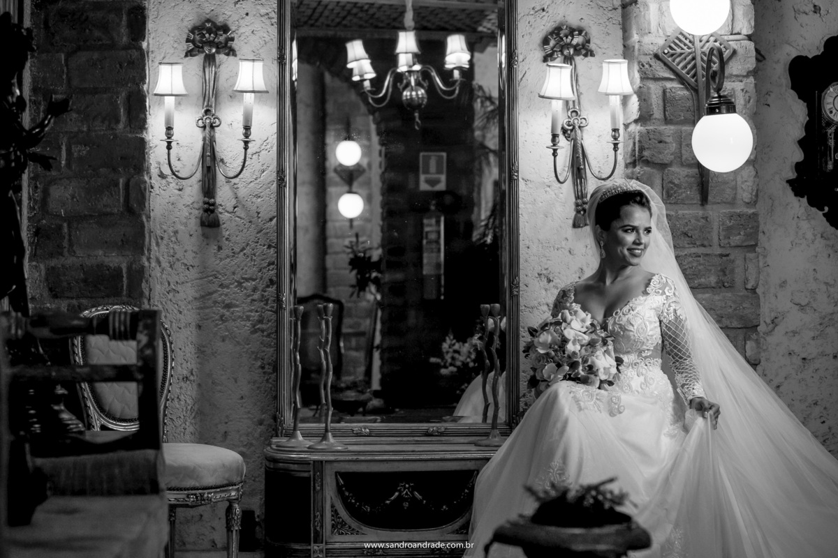 A noiva está sentada de frente para Sandro Andrade seu fotógrafo e segurando o vestido ela olha lateral, linda composição e luz.