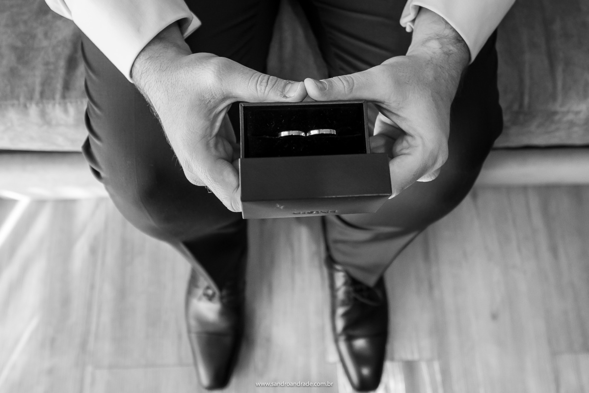 Um ultimo confere nas alianças. Uma fotografia artísticas, feita de cima, onde aparece os pés do noivo, este está sentado segurando as alianças de casamento, Sandro Andrade sempre inovando nos seus clicks e ângulos, fotógrafo premiado do DF.