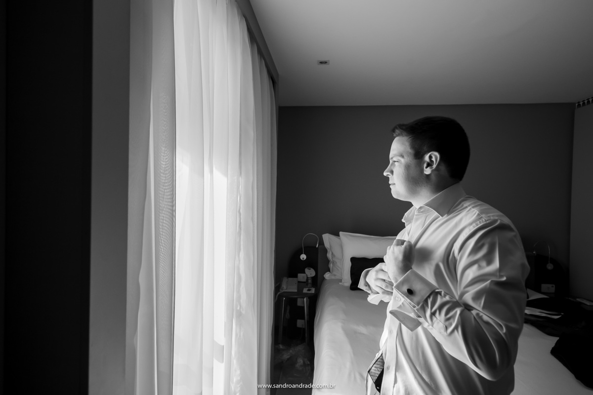 Em uma fotografia preto e branco, o noivo Murilo está de frente para a janela fechando sua camisa.