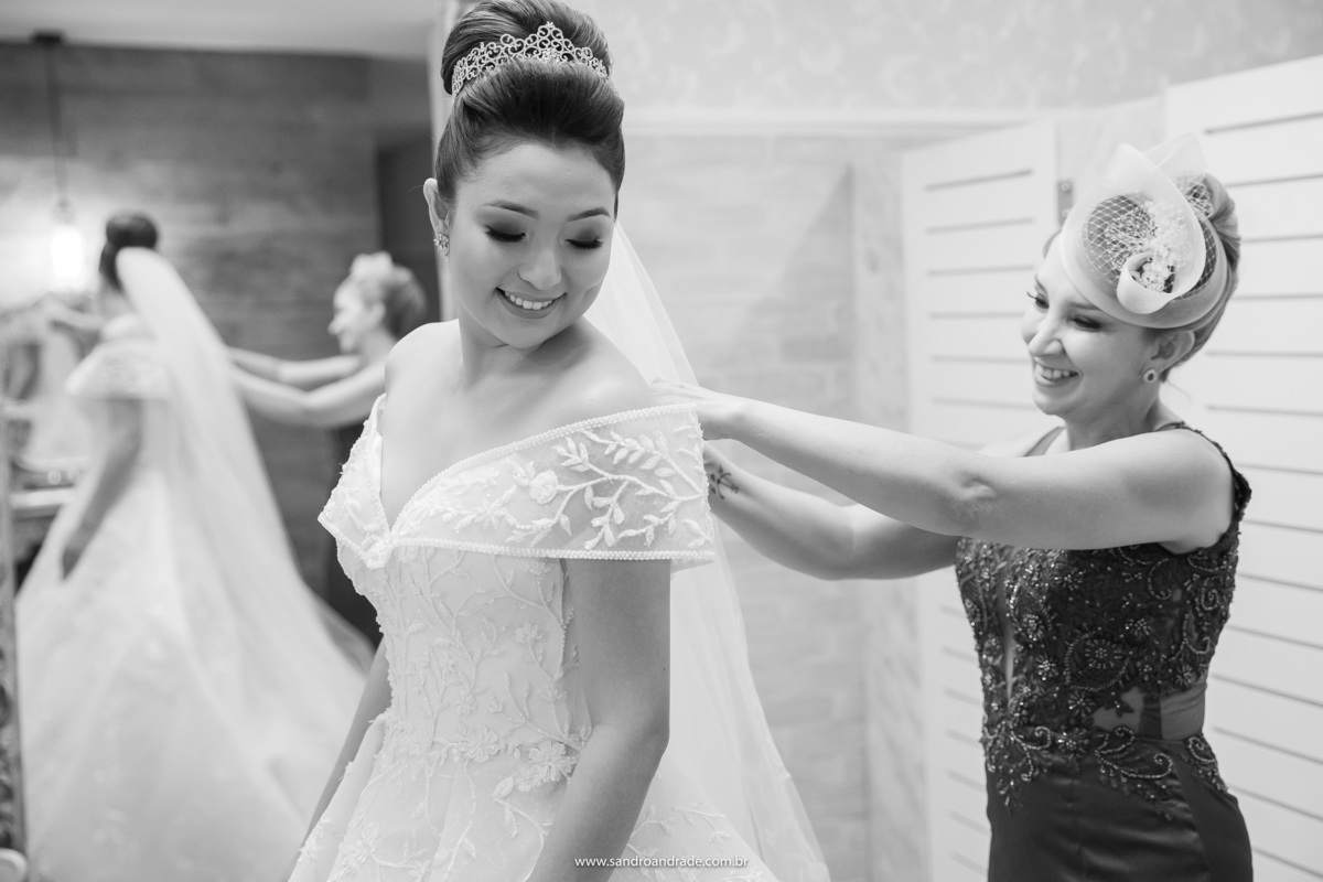 Agora de um angulo diferente, mãe e filha sorriem enquanto fecha o vestido da noiva, sua filha, ao fundo o reflexo das duas no espelho, tudo em uma bela imagem preto e branco.