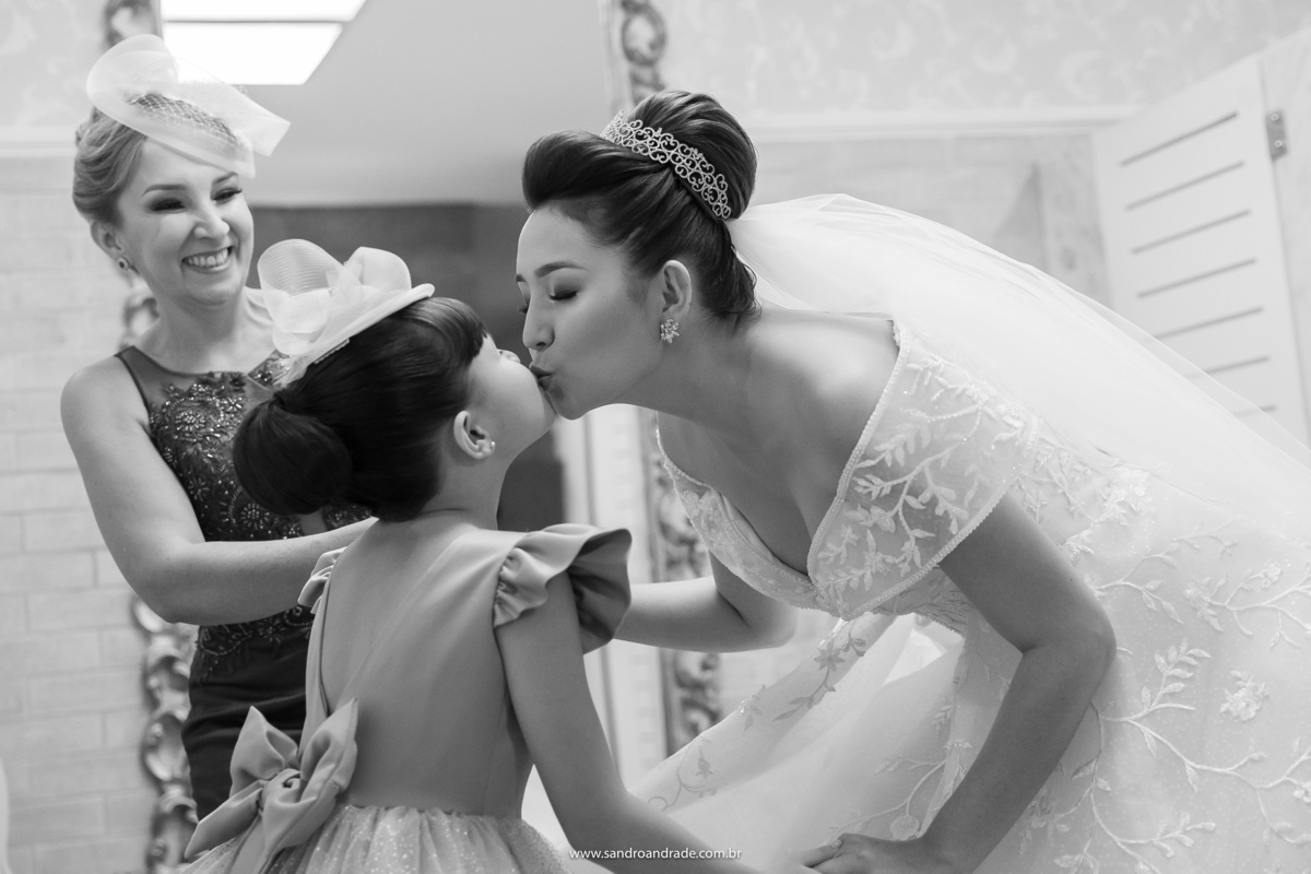 Com a mãe da noiva ao fundo sorrindo, Dany se abaixa para dar um selinho em sua irmãzinha, como ela diz seu bebê. Fotografia de momento por Sandro Andrade um dos melhores fotógrafos de Brasília.
