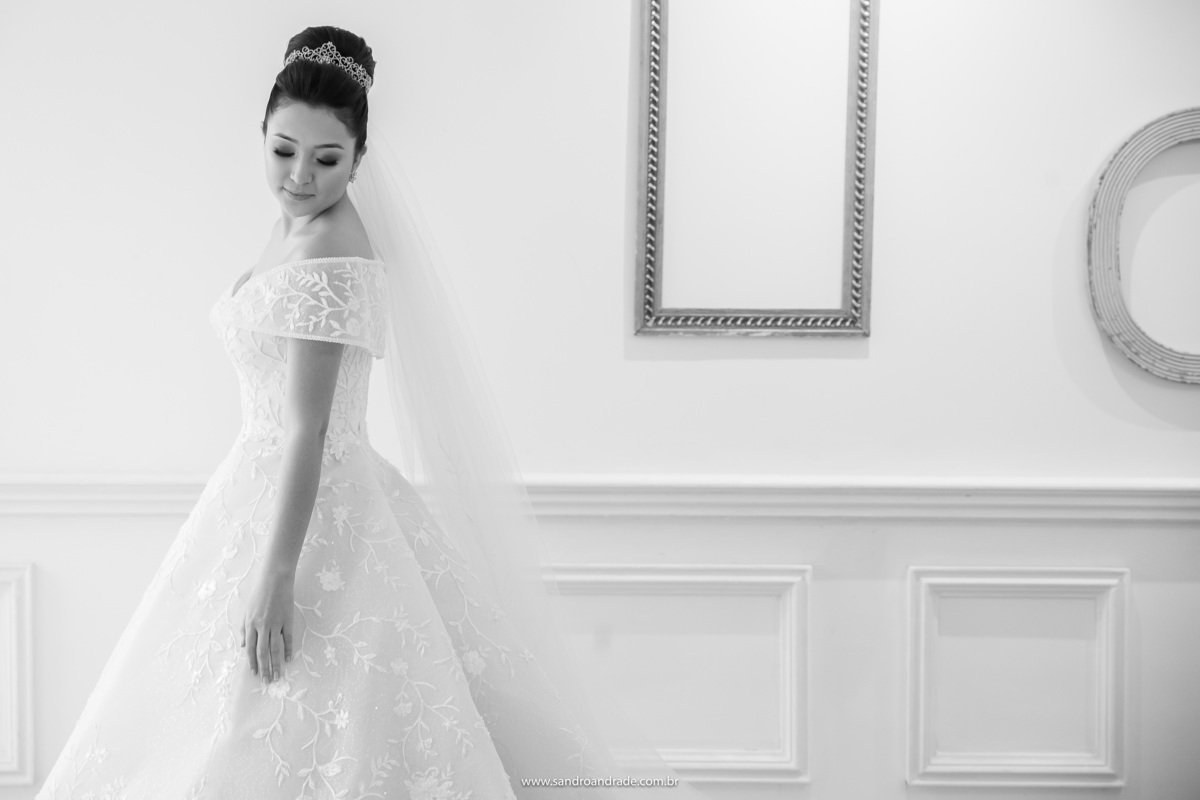 Uma linda fotografia da noiva, em preto e branco, ela esta de lado, olhando a sua cauda do vestido de noiva, ao fundo uma parede branca com molduras.