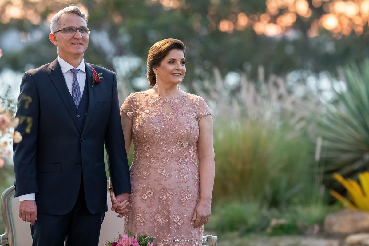 Os pais do noivo de mãos dadas observando a entrada de sua linda nora.