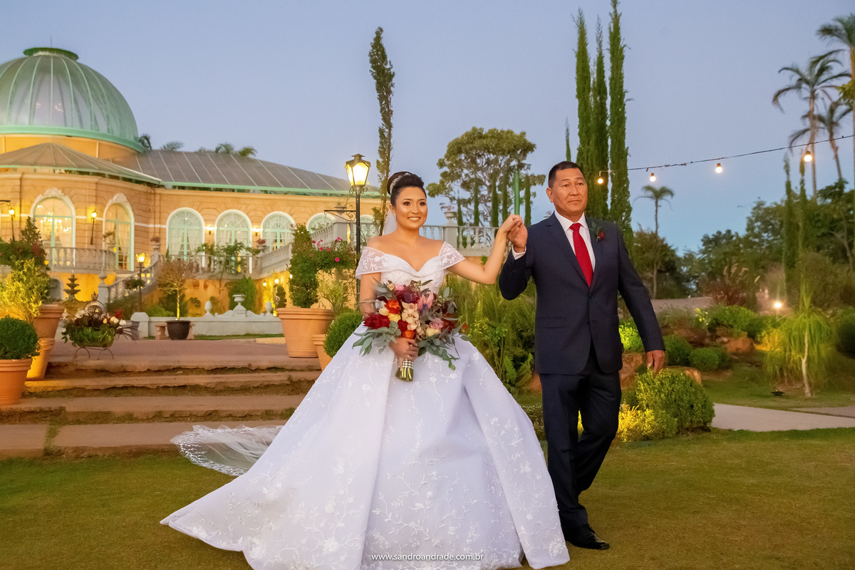 A nossa linda e querida noiva está feliz e sorridente ao lado de seu pai, indo ao encontro de seu amado noivo. Linda fotografia mostrando o palazzo todo iluminado ao fundo e muito verde para todos os lados.
