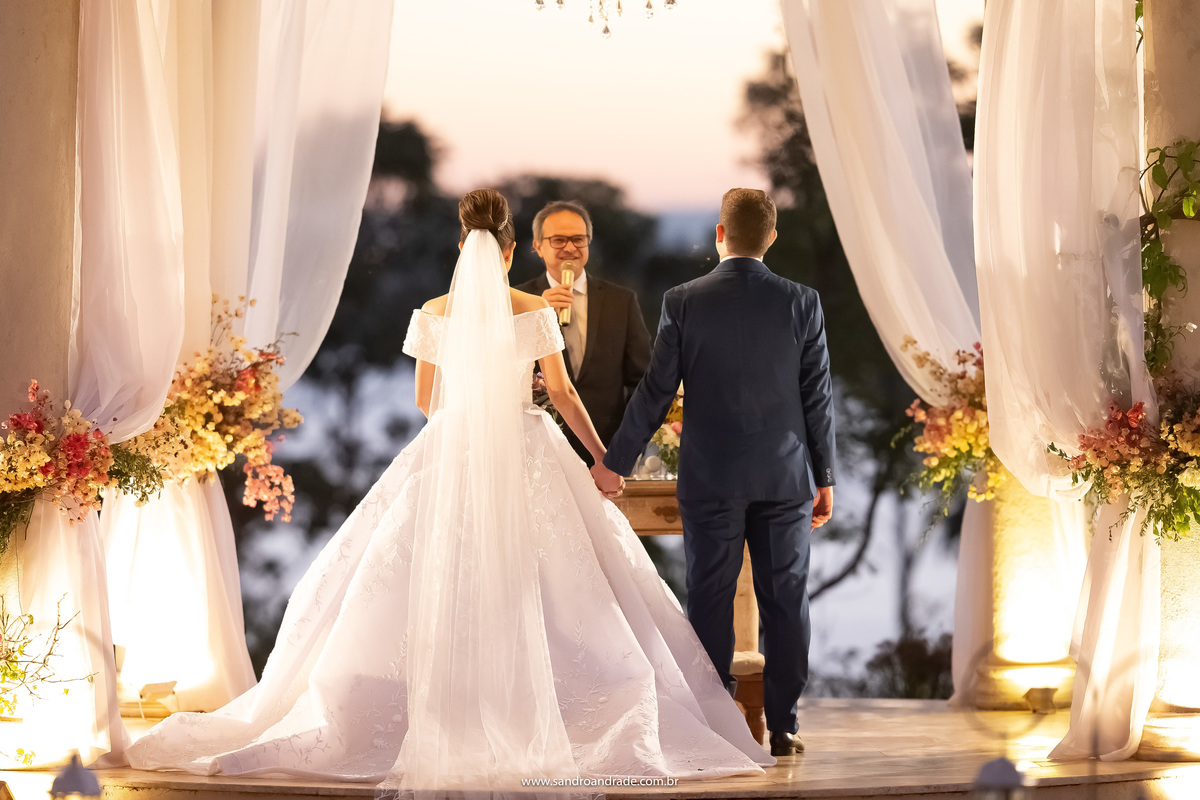 Linda imagem no finalzinho do entardecer, Dany e Murilo estão de costas e o pastor está entre os dois, pegando a vista do lago e as colunas do gazebo iluminado, eles dão inicio a cerimonia de casamento.