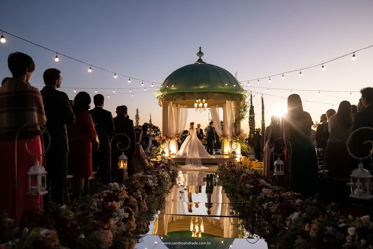 Uma fotografia de trás, pegando o céu, o gazebo, os convidados e o tapete espelhado, o casal está dentro do gazebo e ambos olham para o lado, para olhar para seus convidados, uma fotografia colorida, com um lindo céu em degrade de azul e branco.
