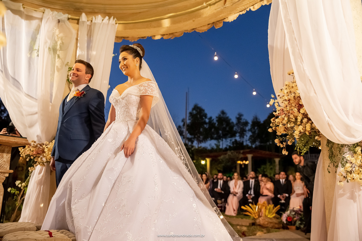 Uma fotografia básica, mas muito bem feita, o casal no altar sorridente, com os convidados ao fundo.