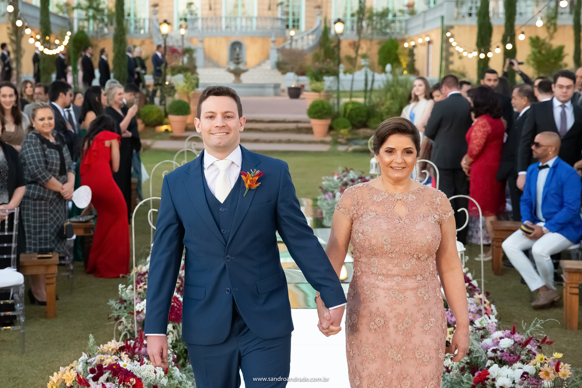 O noivo Murilo a caminho do altar de mãos dadas com sua mãe que está chique demais, uma linda fotografia colorida, mãe e filho, sorrindo e felizes.
