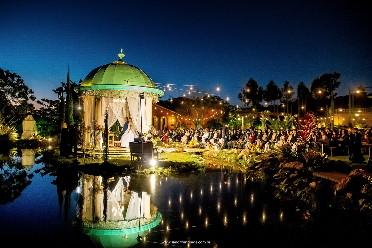 Fotografia colorida, do fotógrafo profissional em Brasilia, BSB ou DF,  com todo o cenário do casamento de Dany e Murilo, o jardim com os convidados, o gazebo com os noivos e o pastor, o lago da Villa com um lindo reflexo e um lindo céu azul ao anoitecer.
