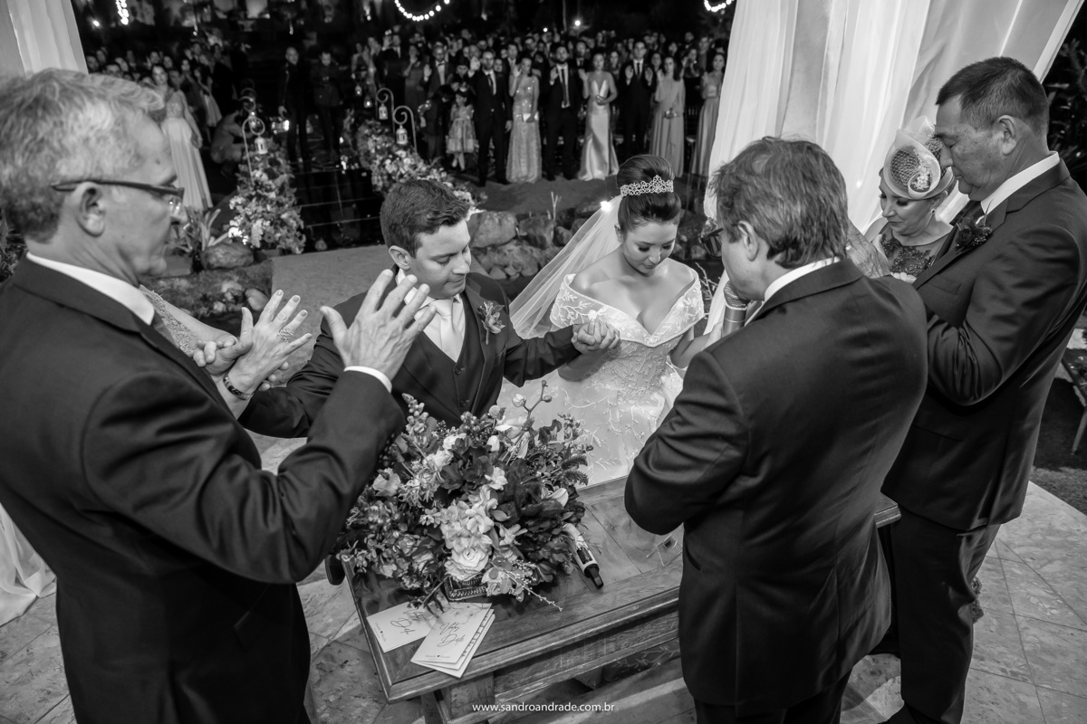 Fotografia preto e branco é sempre muito marcante, dramática, tem toda uma poesia e dessa arte nosso querido Sandro Andrade, mestre em capturar lindos momentos entende muito bem, os noivos estão ajoelhados e com os pais e pastor ao redor, recebem a benção