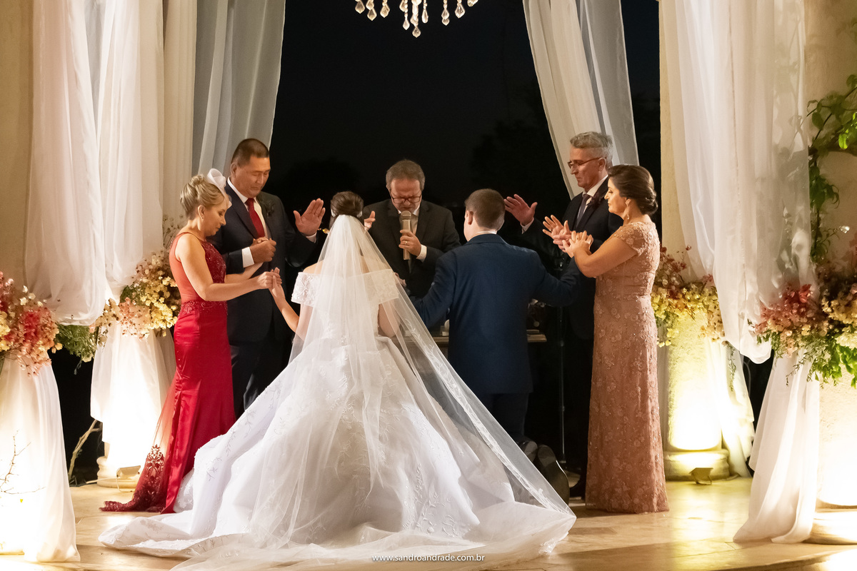 De costas e ajoelhados, os noivos recebem a benção final da cerimonia de casamento, os pais colocam as mãos sobre as cabeças de seus filhos, enquanto as mães dão as mãos a eles. 