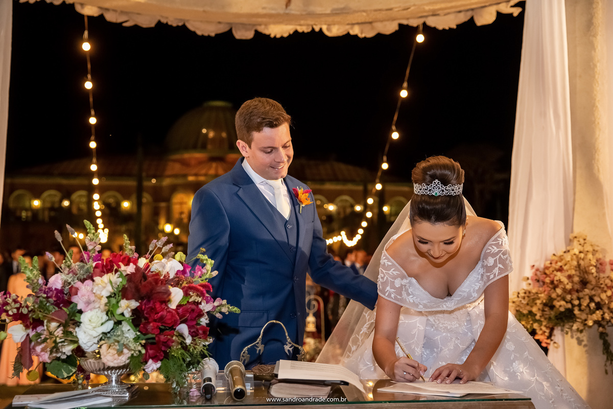 Quase finalizando a cerimonia, Dany está assinando o termo de enlace matrimonial, todo o aplazo aparece ao fundo, Murilo está ao lado dela com a mão em sua cintura.