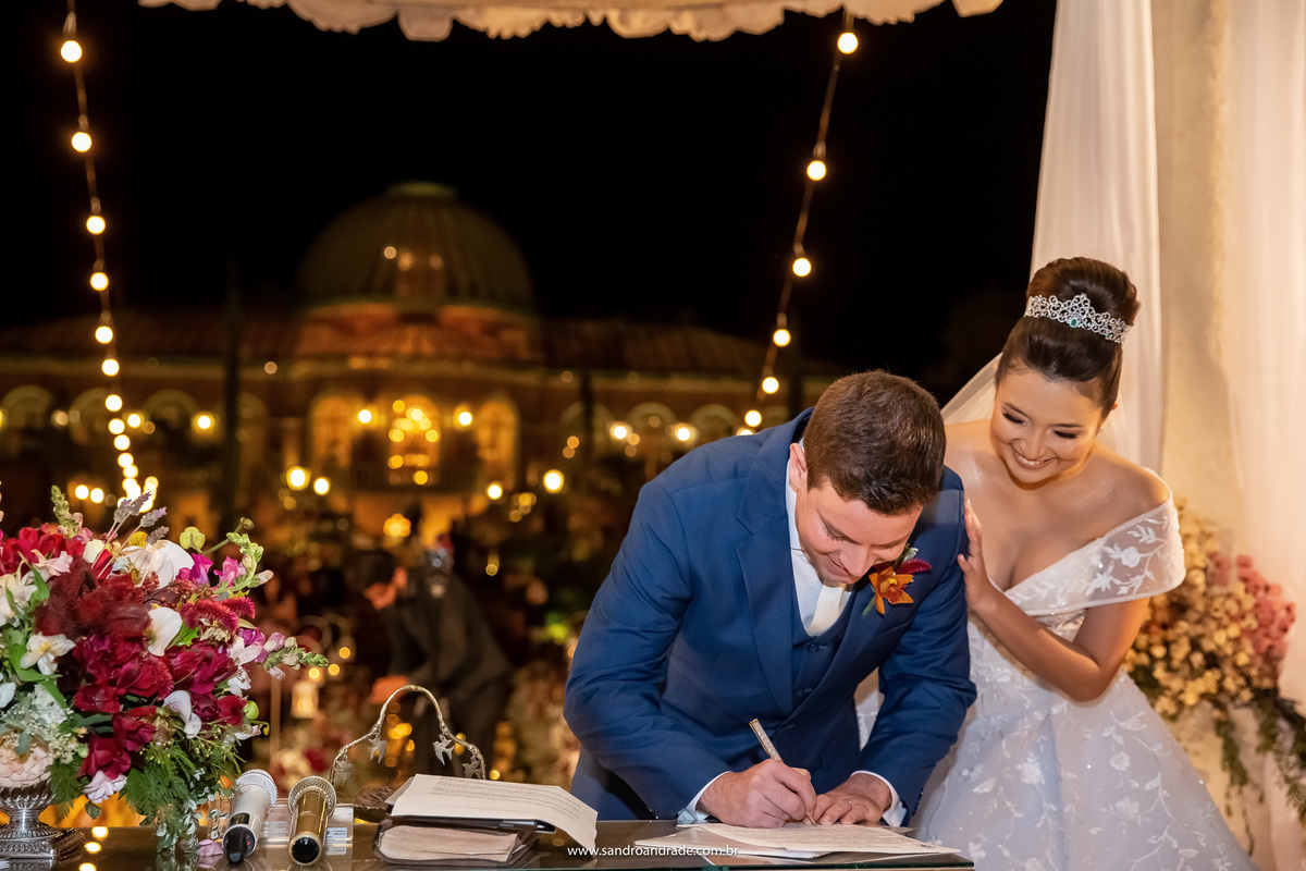 A noiva está abraçada a Murilo que está assinando o termo de enlace matrimonia, os dois sorriem felizes.